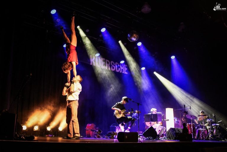 Kuersche Band und Acrobatics Das Bild zeigt den hannoverschen Musiker Kuersche mit seiner Band auf einer Bühne. Eine Akrobatin hält er senkrecht und auf seinen Händen.